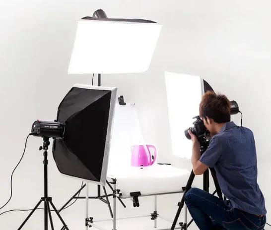 A man is arranging a commercial photo shoot for a kettle on a table in a well-lit shooting room.