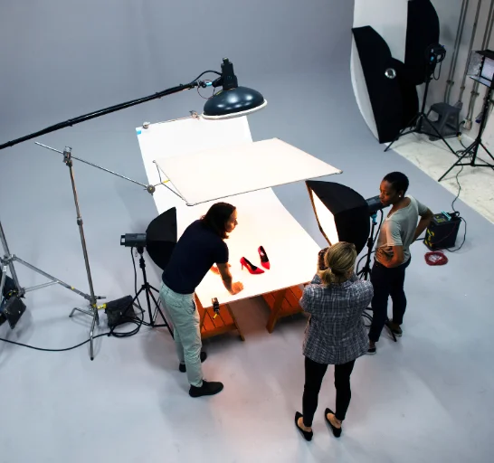 In a well-lit shooting room, three people are setting up a commercial photo shoot for footwear.